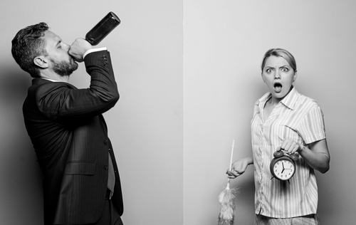 Wife holding a clock and duster in one hand looking shocked at drinking husband.